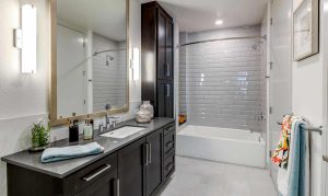 Modern bathroom with dark wood cabinets, a large mirror, gray countertop, and a white bathtub surrounded by glossy subway tiles.